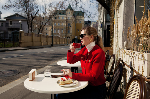 Portrait beautiful tourist woman drinks cup coffee and eats cake, sits in street cafe in city center. Elegant girl wears red blazer jacket, white turtleneck and sunglasses