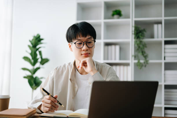 istock Male student taking notes from a book at library, Young asian sitting at desk doing assignments in college library 1483503665
