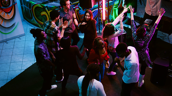 Multiethnic group of friends partying, showing funky dance moves at nightclub. Men and women dancing on electronic beats having fun on night out, enjoying colorful spotlights. Handheld shot.