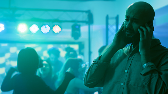 Male adult chatting on phone at party, trying to talk to someone on mobile phone remote call in discotheque. Young person attending dance party at club, answering smartphone. Tripod shot.