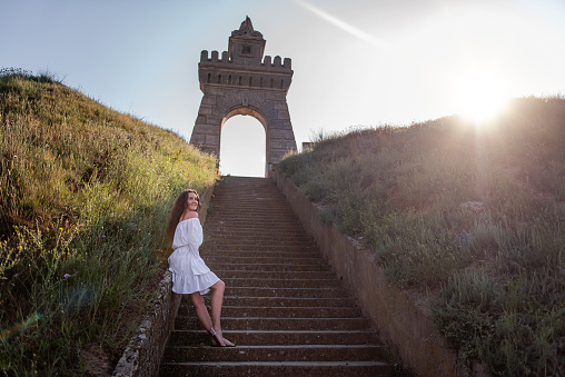 Young, happy woman dancing in white, summer dress stands on concrete staircase among the green hills against the backdrop of ancient architecture, vintage arch at sunset. Girl travels on the weekend