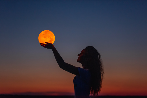 A young woman holds the full moon in her hands against the backdrop of a red sunset. Millennial blogger leads social media. Girl silhouette portrait. Astrology, Waxing Crescent. Copy space