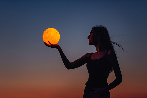 A young woman holds the full moon in her hands against the backdrop of a red sunset. Astrology
