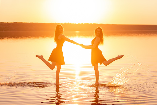 Two sisters in black dresses dance on the water, holding hands in the contrasting rays of the sunset. Girls are having fun on the journey, circling together. Girlfriends rest on the sea on a travel