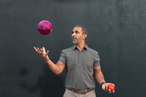 A male juggling a cabbage and a tomato against a black background