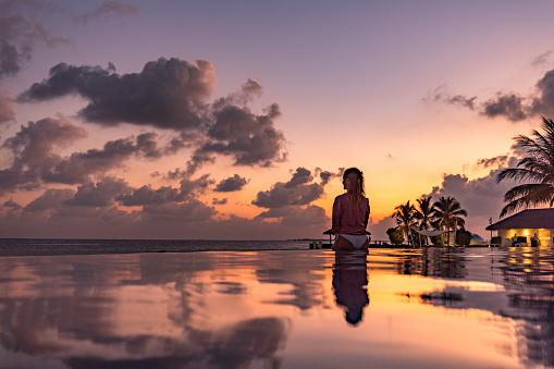 Back view of a woman relaxing at the edge of an infinity pool at sunset. Copy space.
