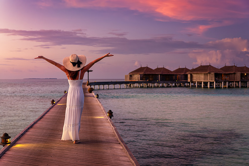 A happy woman in white dress and hat walks down a pier over turquoise ocean in the Maldives islands during colorful sunset time