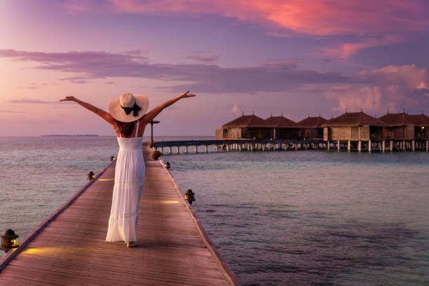 a woman in white dress walks down a pier over turquoise ocean in the maldives during sunset - hotel tourist resort luxury tropical climate imagens e fotografias de stock