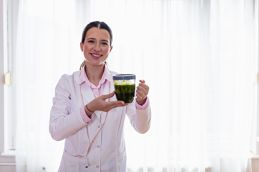 A young female nutritionist is shown holding a glass of fresh green smoothie in her hand