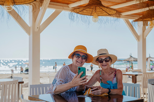 Two older women in hats are using cell phones and smiling at a beach bar by the sea. Summer vacation concept.