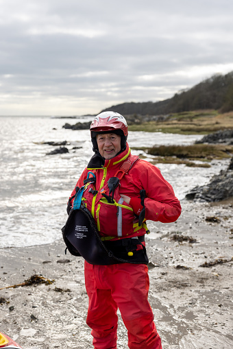 A front view full length of a kayak team member who is proud to be part of team he is holding his arms in his lifejacket and looking into the camera. He is also wearing helmet and a dry suit. He is also on the beach in Dumfries.
