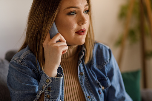 Portrait of concerned young woman sitting on the sofa, looking away, contemplating while having a feud with a friend over the phone.