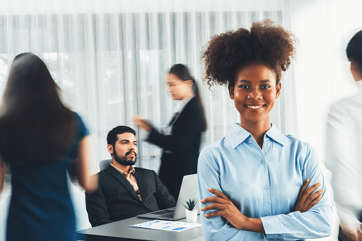 Young African businesswoman portrait poses confidently with diverse coworkers in busy meeting room in motion blurred background. Multicultural team works together for business success. Concord