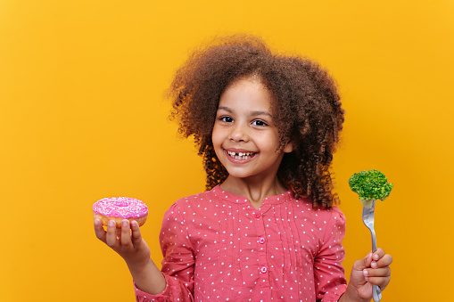 Studio shot of a smiling Black girl holding fresh broccoli on the fork on yellow background. The concept of choosing products for healthy baby food.