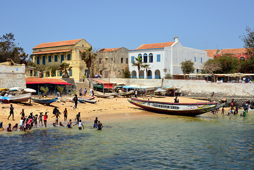 Gorée Island, Dakar, Senegal: the 'Petite Plage', small beach with golden sand, protected from the waves of the Atlantic by the harbor breakwaters - boats on the sand and buildings in Goverment Square / Hesse Street - Maritime Museum ('Musée de la Mer') and Dumont house, with a ruined building in between - East coast of Gorée Island - UNESCO world heritage site.