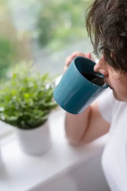 A woman in white t-shirt is drinking coffee at home in front of window and looking outside.