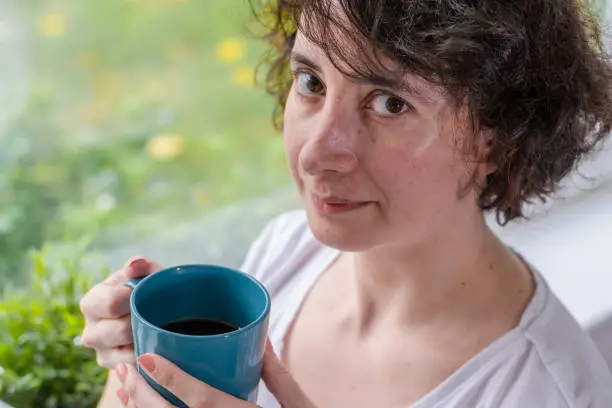 A woman is holding a coffee mug at home, in front of a window and looking at the camera, smiling, at her 40s and wearing a white t-shirt.