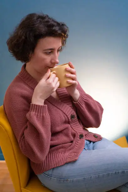 Woman at her 40s is sitting on a yellow chair at home and drinking coffee or tea from a yellow mug, looking thoughtful.