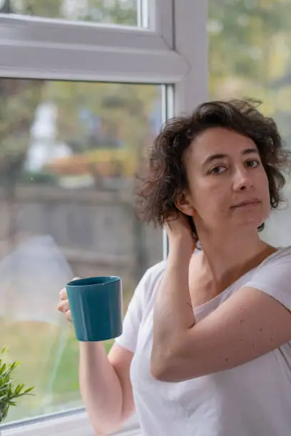 Woman fixing her hair in front of a window, wearing a white t-shirt and holding a dark green mug on one hand.