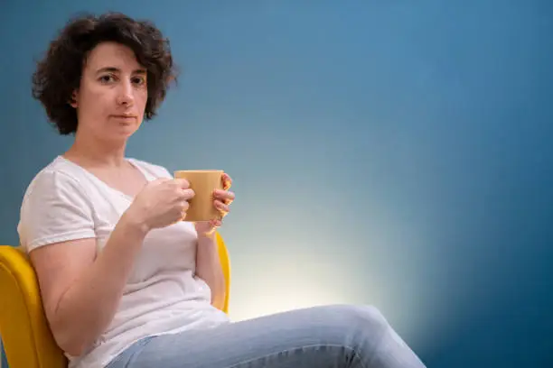 Woman holding a yellow mug, looking at the camera, wearing a white t-shirt and a pair of jeans, sitting on a yellow chair in her living room in front of a dark blue wall. She’s around 40 years old.