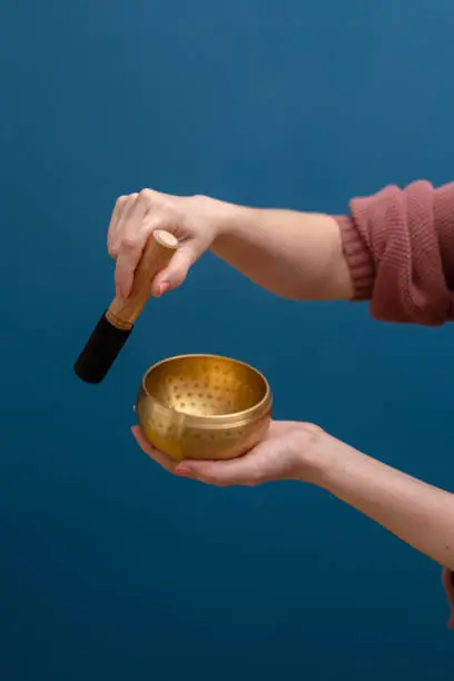 A woman’s hands on a vertical image, holding a yellow zinc metal Tibetan bowl on one hand and using the playing stick, in front of a dark blue wall.