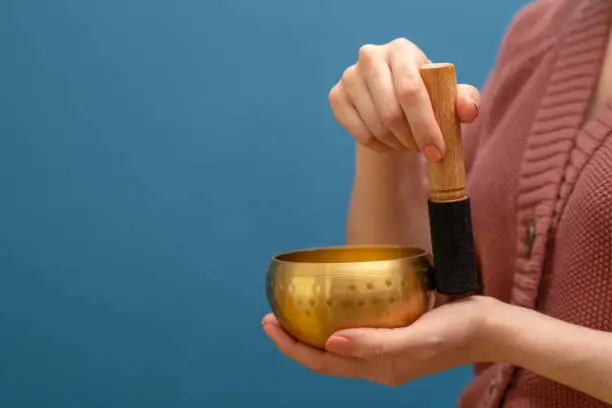 Vertical image of a woman’s hands holding a yellow coloured zinc Tibetan singing bowl in front of a dark blue wall and using it.