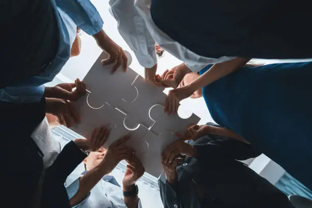 Below view of diverse corporate officer workers collaborate in office connecting puzzle pieces as partnership and teamwork concept. Unity and synergy in business idea by merging jigsaw puzzle. Concord