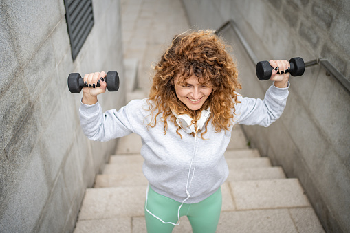 A woman trains outdoors with dumbbells during the day, she is in a part of her apartment complex, next to a concrete wall, where she regularly exercises