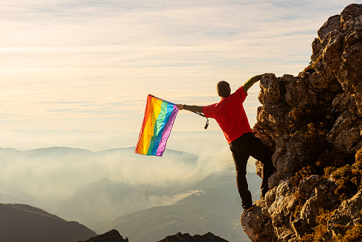 mountaineer man making summit on a mountain and waving a rainbow lgbt pride flag at sunset. lgbt movement and rights concept . Concept of conquering rights of lgbt people.