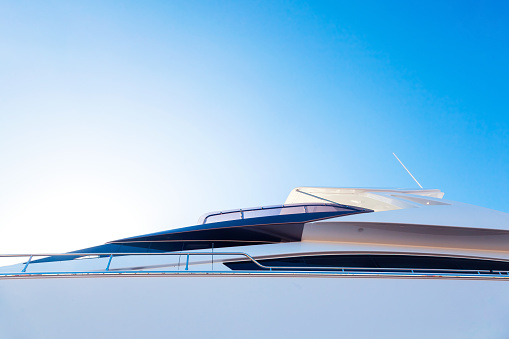 The hull of a luxury, motor, white yacht, with portholes on it, against the blue sky and the light of the sun in the corner, bottom view.