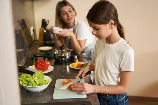 Teenager girl is cuting tomatoes for breakfast in the kitchen. Her mother is standing nearby, drinking coffee and giving advice to the child. Plates with food are on the kitchen surface