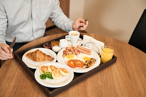 Tasty breakfast of different blues is standing on the table. Croissant, cheese, sausages, bacon and eggs, fried mushrooms, orange juice, hot coffee with milk and sugar. Breakfast concept in hotel