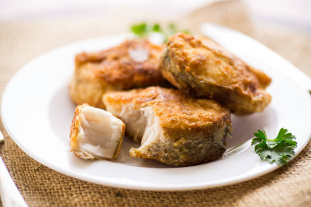 A piece of hake fish fried in a crispy crust, in a plate on a wooden table.