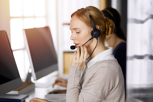 Businesswoman talking by headset while sitting in sunny office. Call center and diverse people group in business.