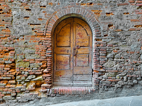Old closed italian traditional wooden door against an old brick wall with arched opening - Tuscany - Italy