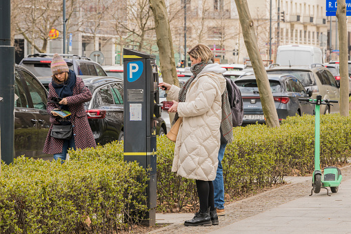 Side view of a woman taking a parking ticket and paying on a parking vending machine in the street during day time. 5 april 2023, Warsaw, Poland.