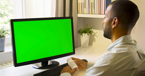 Side view of man holding pills bottle while using green screen of computer monitor in home office.