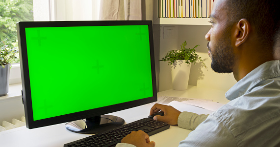 Side view of man working at green screen of computer monitor in home office.