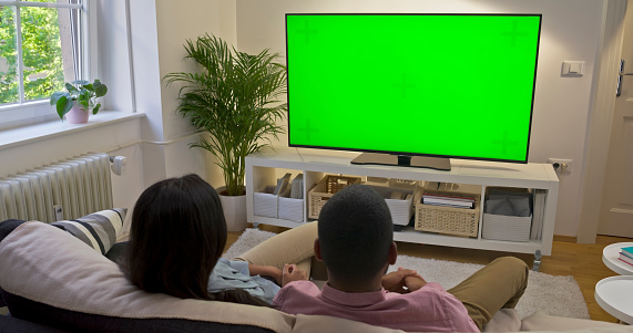 Rear view of couple watching green screen TV while sitting on sofa in living room.