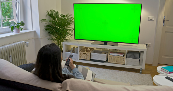 Woman watching green screen TV while sitting on sofa in living room.