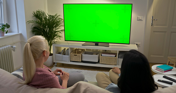 Women watching green screen TV while sitting on sofa in living room.