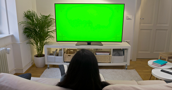 Rear view of woman watching green screen TV while sitting on sofa in living room.