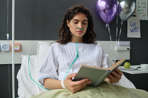 Young girl enjoying reading of book while lying on bed in hospital ward