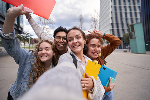 Smiling multiracial college students taking a selfie back to school. Happy friends photo together outside campus university holding folders. Cheerful classmates young people pose and amused.