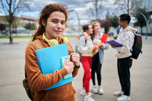 Red-haired college boy poses smiling looking at camera outside. Young millennial cheerful student holding folder of notes with happy classmates in background. Life on campus during exam time.