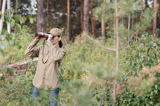 A woman forester in uniform looks through binoculars and monitoring the forest area in summer, selective focus. Ecologist, biologist, park ranger, nature, environmental conservation concept
