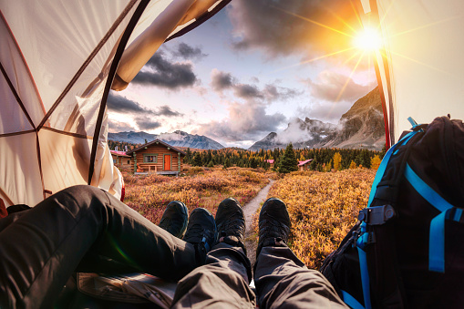 Legs of couple hiker relaxing inside a tent with Rocky Mountains and wooden hut on autumn forest in national park at the sunset