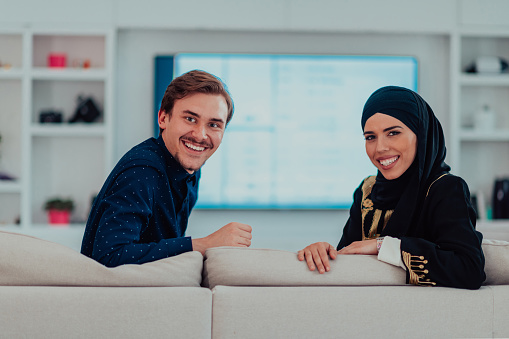 Young muslim couple woman wearing islamic hijab clothes sitting on sofa watching TV together during the month of Ramadan at modern home.