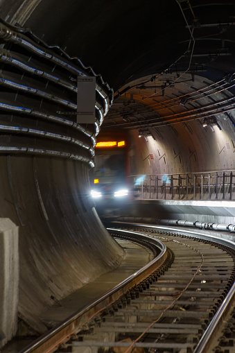 A muni train is coming into a tunnel. The train has its lights on and only the first part of the train can be see. The tunnel is lit and pipes and tracks can be seen.