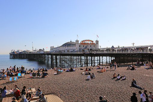 Brighton, United Kingdom - April 15, 2022: Brighton Palace Pier with people sitting on the beach, East Sussex, England.